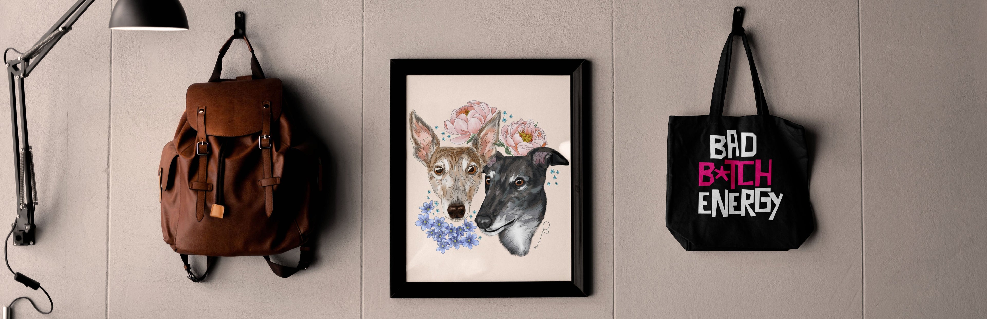 A bare panel wall with a framed digital artwork of two greyhound dogs with flowers around them (one brindle dog, one black dog, flowers are forget me not and peonies, on a light pink background), a brown backpack, and a black tote bag with printed grunge text in white and pink that says "bad b*tch energy"