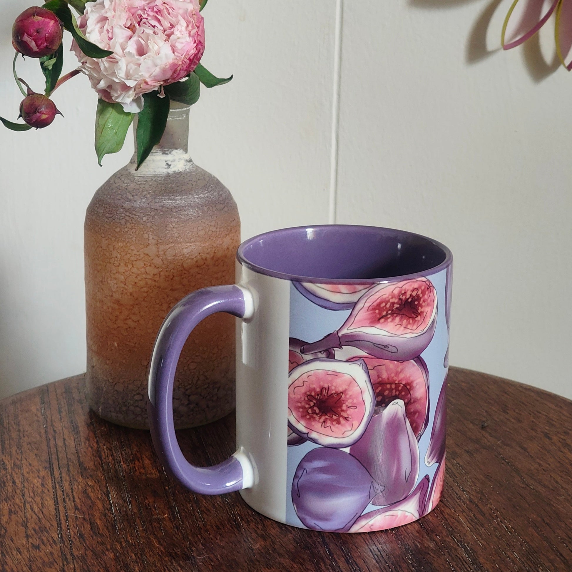 Mug with fig design next to a vase with pink flowers on a wooden surface.