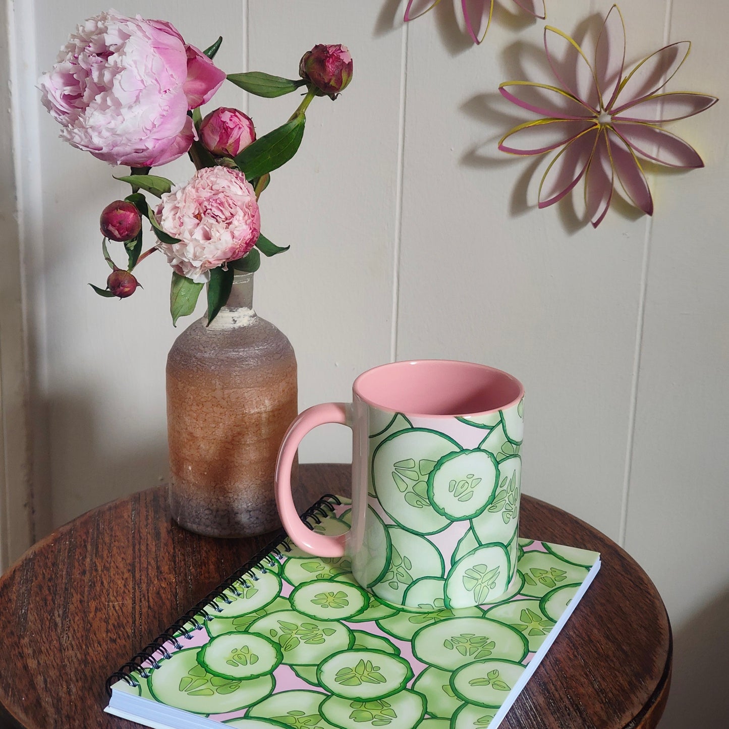 Pink mug with cucumber design on a table with flowers and a notebook.