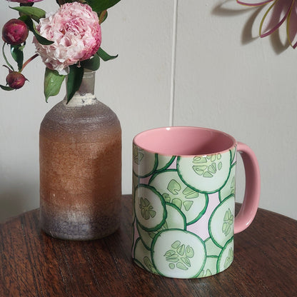 Vase with flowers and a mug witch cucumber print on the outside, with a pink handle and interior. It sits on a wooden surface with decorative wall art in the background.