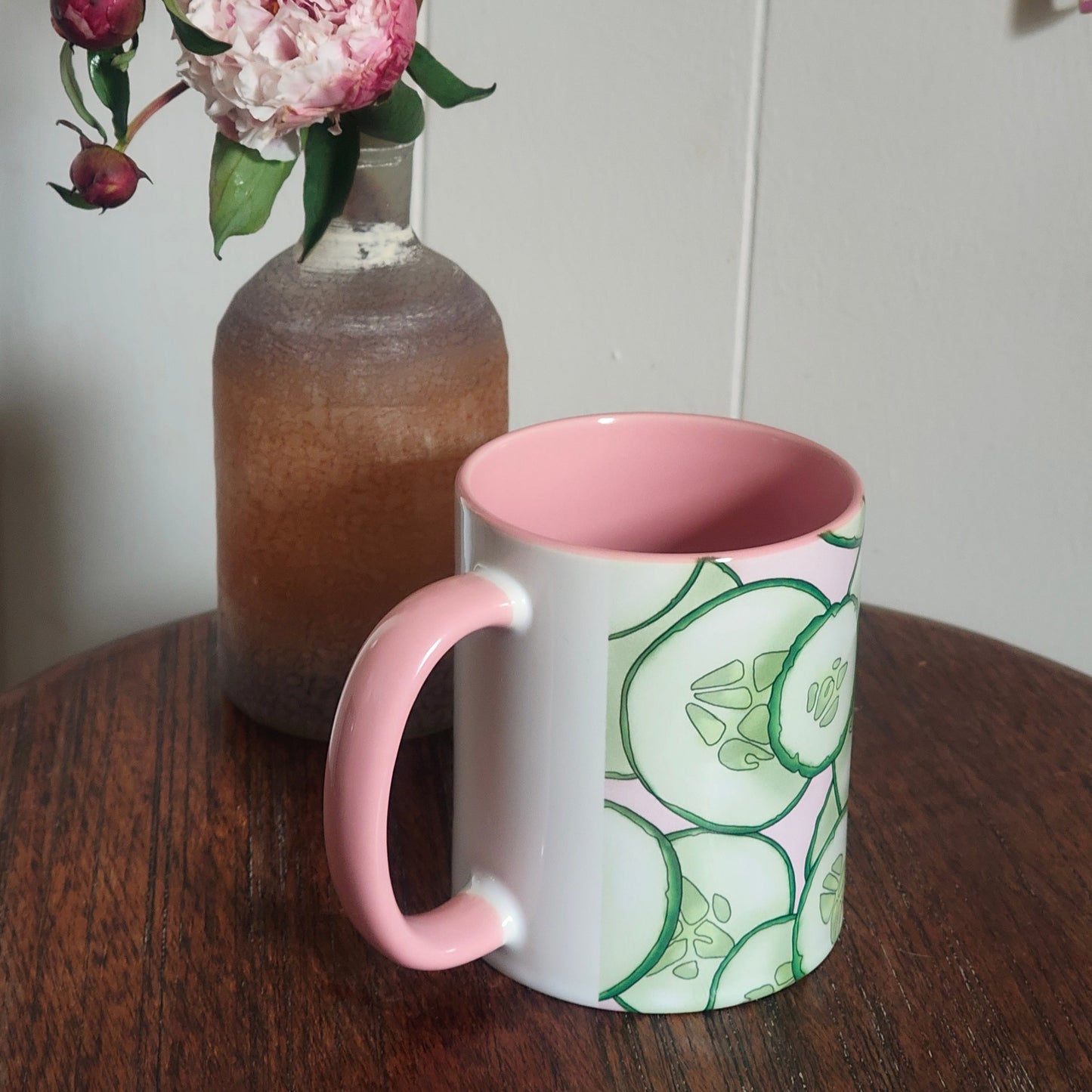 Mug with pink interior and handle, cucumber design on exterior, next to a vase with flowers on a wooden surface.