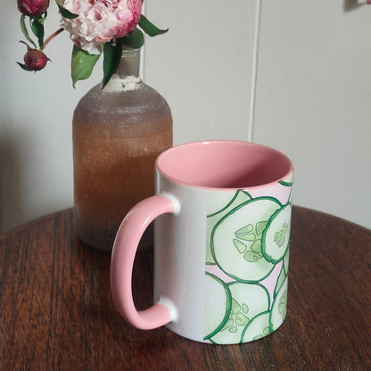 Mug with pink interior and handle, cucumber design on exterior, next to a vase with flowers on a wooden surface.