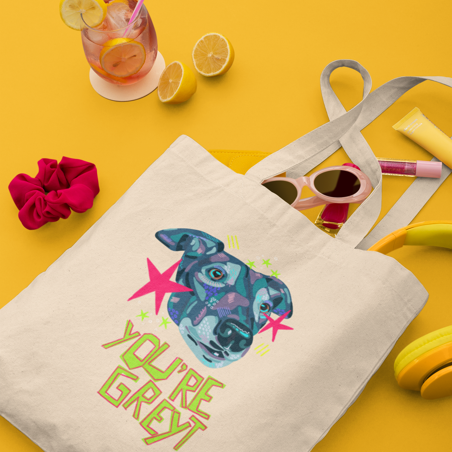 A cream canvas tote bag with a colorful graphic of a dog's face and the text 'YOU'RE GREY' in green, styled on a bright yellow background with sunglasses, headphones, a drink, and a pink scrunchie.