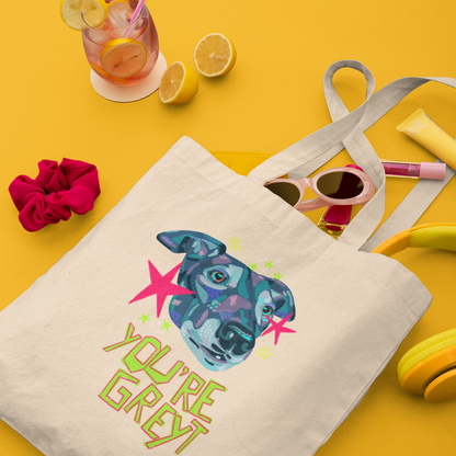 A cream canvas tote bag with a colorful graphic of a dog's face and the text 'YOU'RE GREY' in green, styled on a bright yellow background with sunglasses, headphones, a drink, and a pink scrunchie.