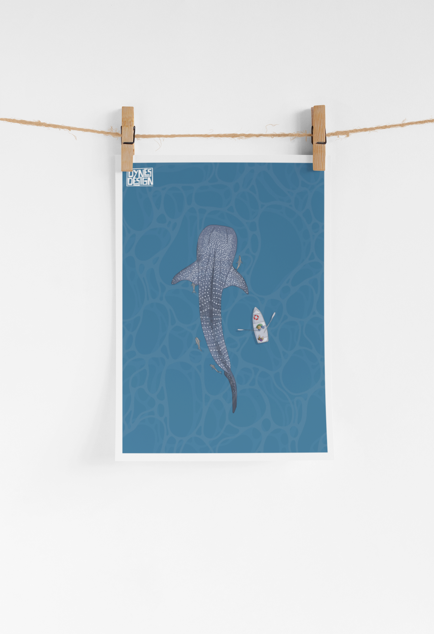A vertical art print featuring a whale shark and a small boat on a blue water background, hanging from a string with two wooden clothespins against a white wall.