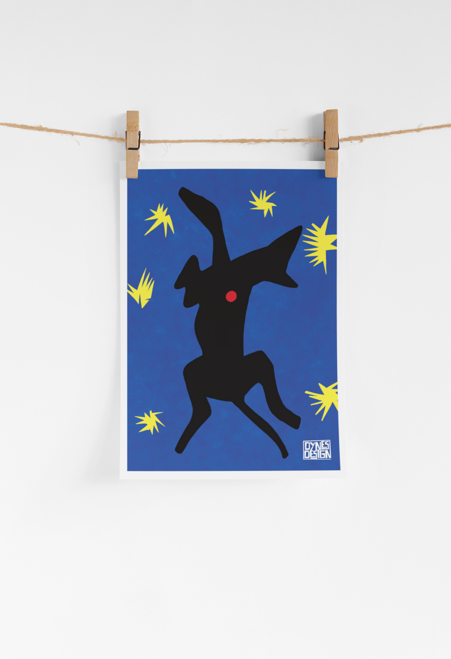 A vertical art print featuring a black greyhound silhouette with a red dot on a blue background, decorated with yellow starburst shapes. The print is hung from a wooden clothesline with two wooden clothespins against a clean white wall.