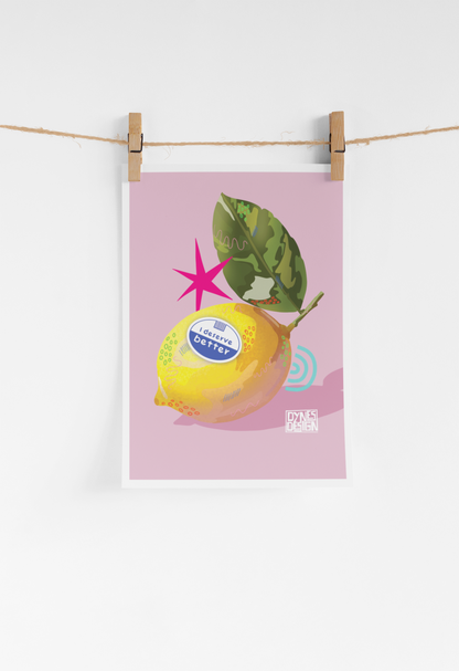 A vertical art print featuring a stylized yellow lemon with a green leaf on a dusty pink background, hanging from a string with two wooden clothespins against a white wall.