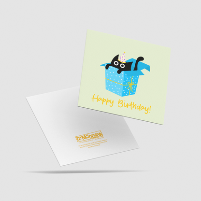 A square birthday card with another behind showing the back, shown on a light gray background. The card features a pale yellow-green background with a cartoon black cat peeking out of a blue gift box, and the words 'Happy Birthday!' in yellow.
