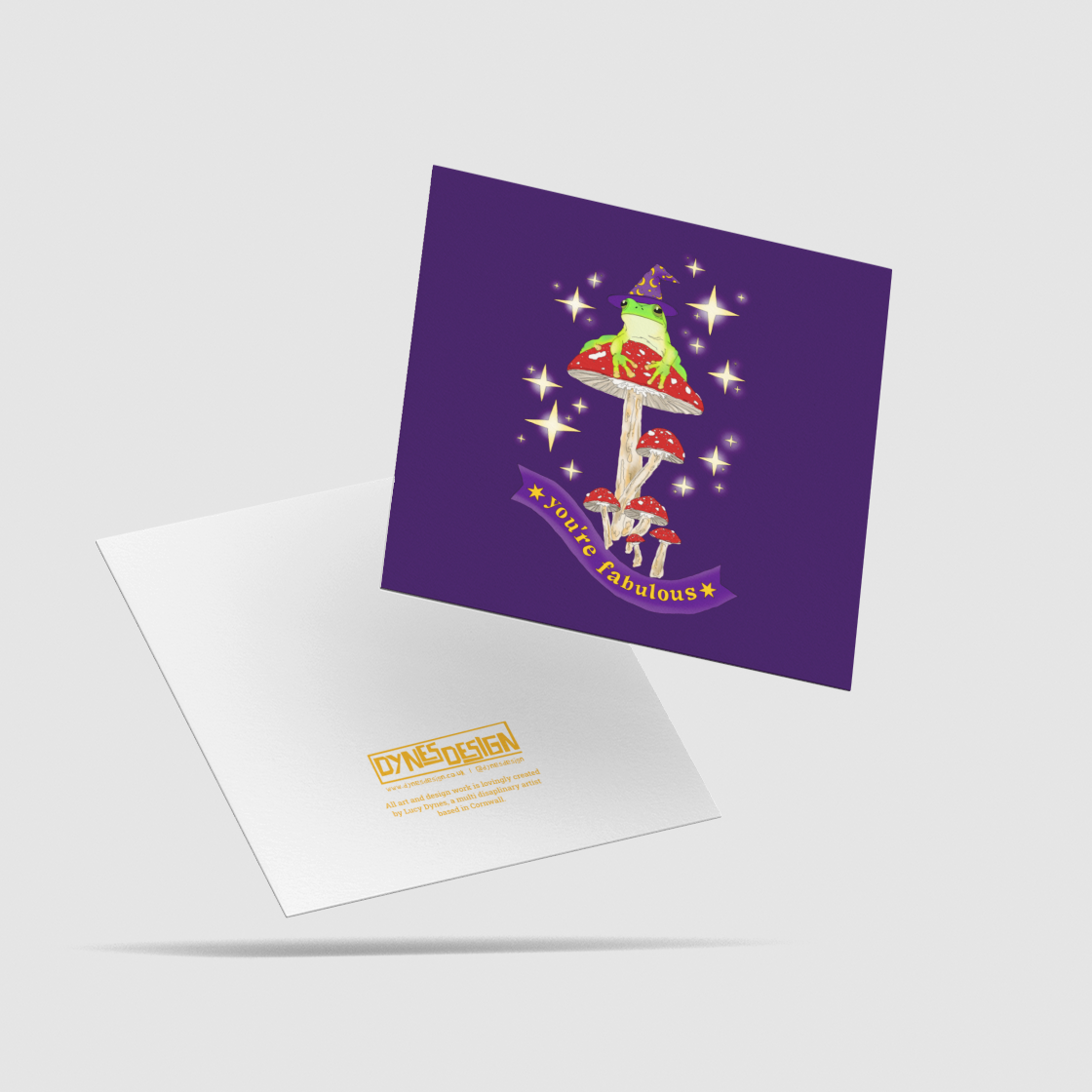 A square purple greeting card with a colorful graphic of a wizard frog on-top of a cluster of mushrooms, surrounded by glittering stars, with a banner that says, 'you're fabulous" on it. Behind is the back of the card with the logo mark "DYNES DESIGN"