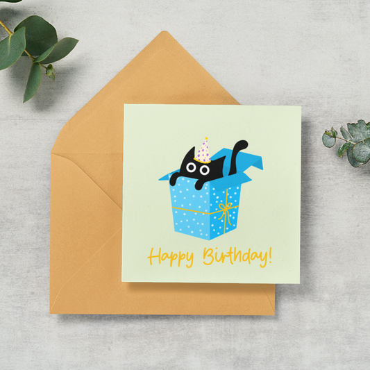 A square birthday card with a cartoon black cat popping out of a blue gift box, placed on top of a tan envelope on a light gray textured surface with green eucalyptus leaves around it.