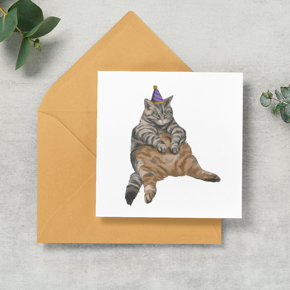 A square white greeting card with a watercolor illustration of a tabby cat wearing a party hat, placed on top of a tan envelope on a light gray textured surface with eucalyptus leaves around it.