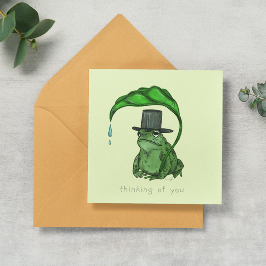 A square greeting card with a light yellow-green background, featuring an illustration of a green frog wearing a black top hat under a large green leaf, placed on top of a tan envelope. The card reads 'thinking of you' and is styled on a light gray textured surface with eucalyptus leaves.