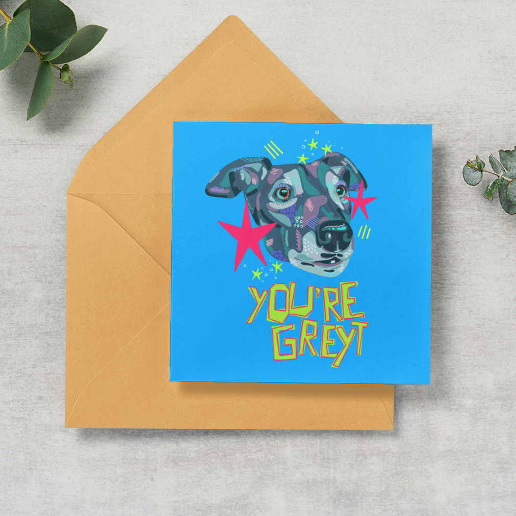 A square blue greeting card with a colorful dog illustration and the words 'YOU'RE GREY!' in yellow, placed on top of a tan envelope on a light textured surface with green eucalyptus leaves around it.