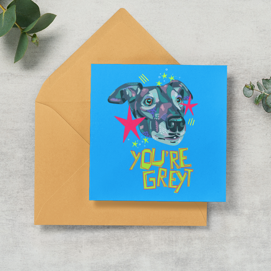 A square blue greeting card with a colorful dog illustration and the words 'YOU'RE GREY!' in yellow, placed on top of a tan envelope on a light textured surface with green eucalyptus leaves around it.