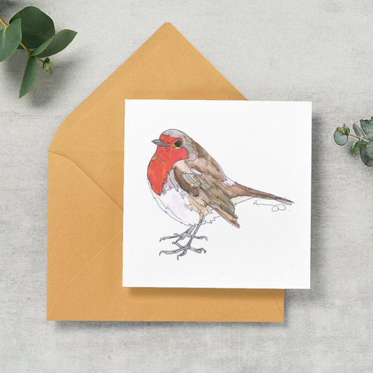 A square white greeting card featuring a watercolor illustration of a robin bird, placed on top of a tan envelope on a light gray textured surface with green eucalyptus leaves around it.