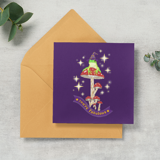 A square purple greeting card with a whimsical mushroom illustration and a natural kraft envelope, styled on a light gray textured surface with green eucalyptus leaves.
